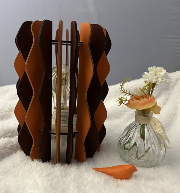 Lampe Pinus en bois à lamelles alternant deux couleurs (Orange et marron). Un oiseau en 3D et un pot de fleurs sont à coté de cette lampe, le tout sur une nappe.